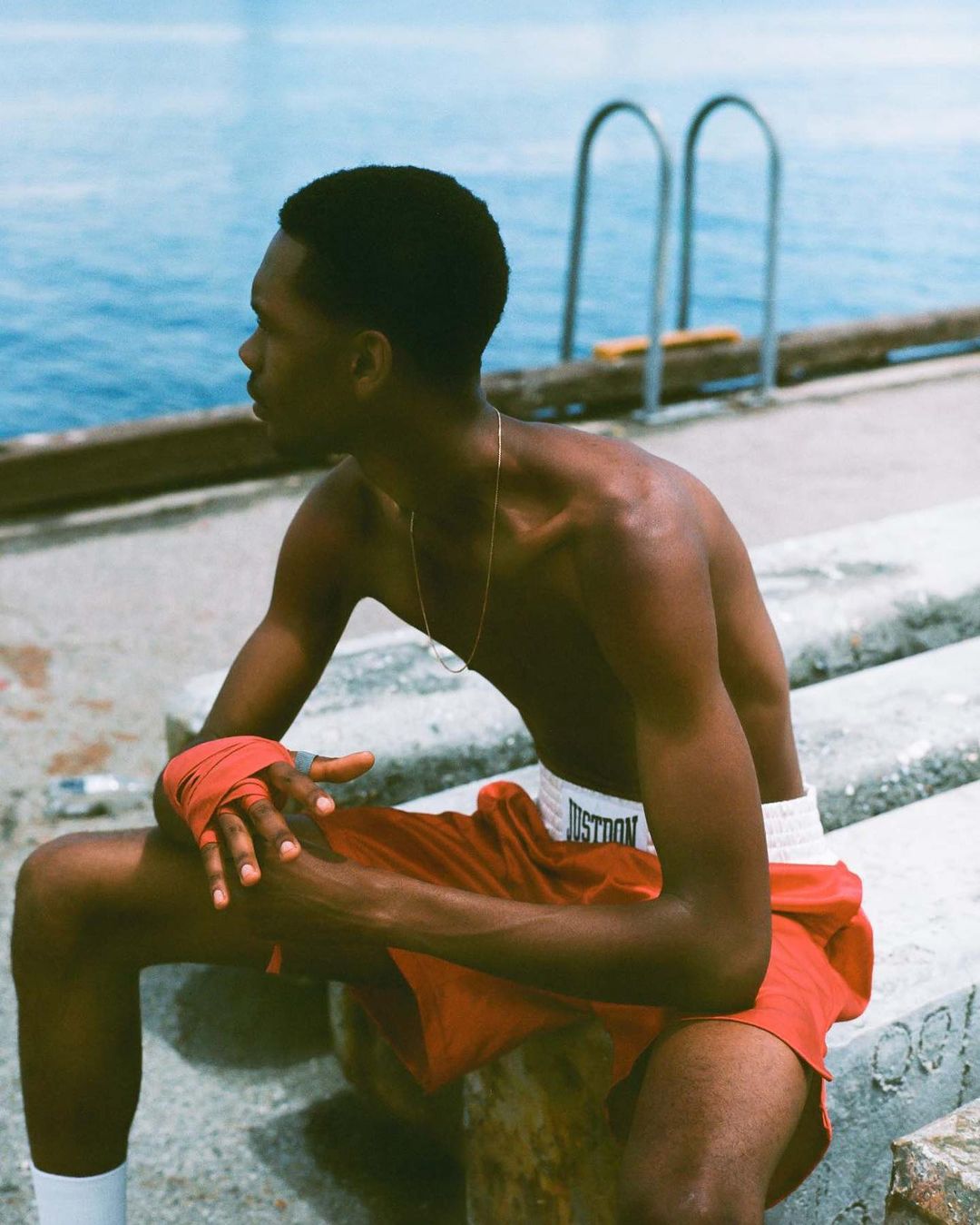 Shirtless man with hand wraps and boxing shorts sits along a waterfront, looking at the blue ocean with a ladder in the distance.