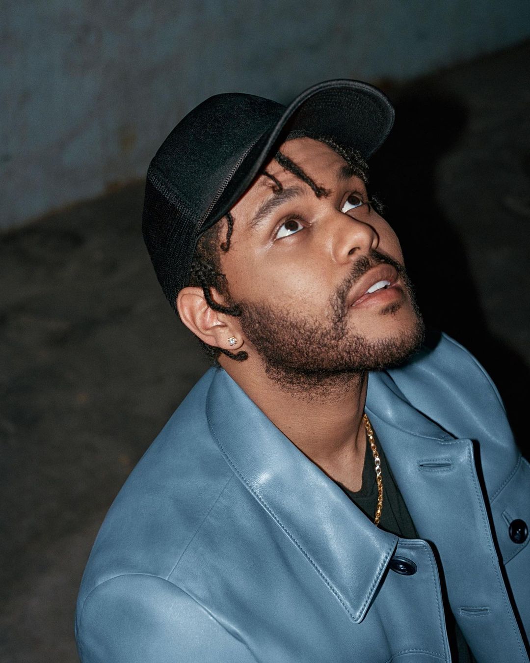 Portrait of a stylish adult male in a blue jacket and black cap looking upward with a neutral expression in a dimly lit space.