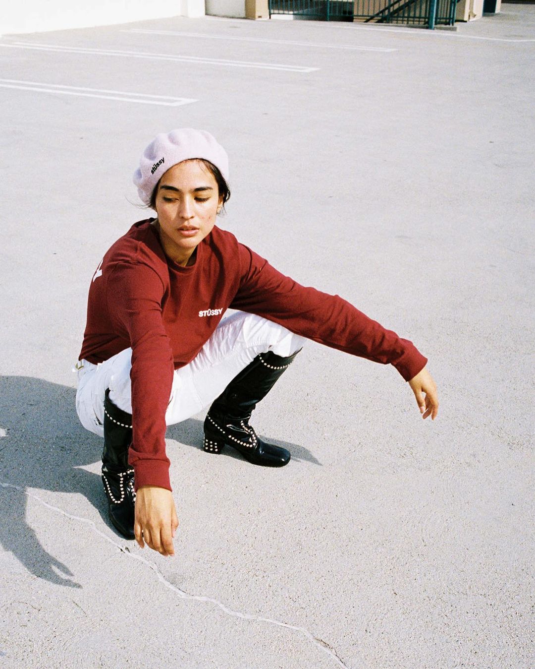 An adult female squats on concrete wearing a red long-sleeved shirt, white pants, black boots, and a light-purple beret in daylight.