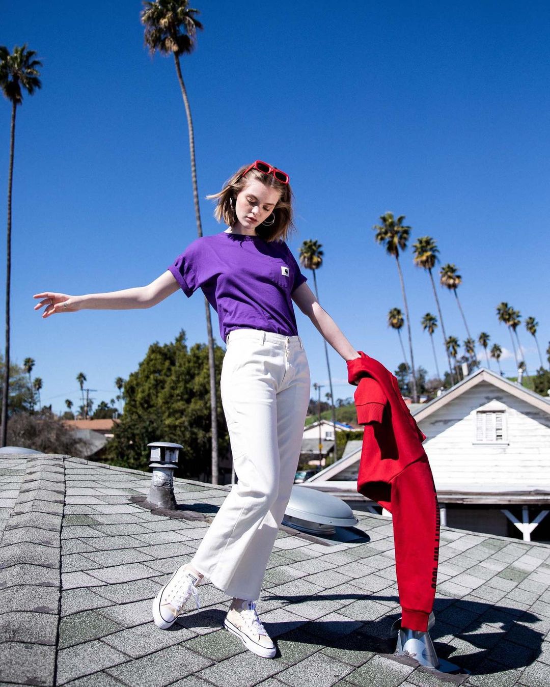 Woman in purple tee and white jeans poses on a rooftop with palm trees and a blue sky, holding a red jacket and wearing red sunglasses.