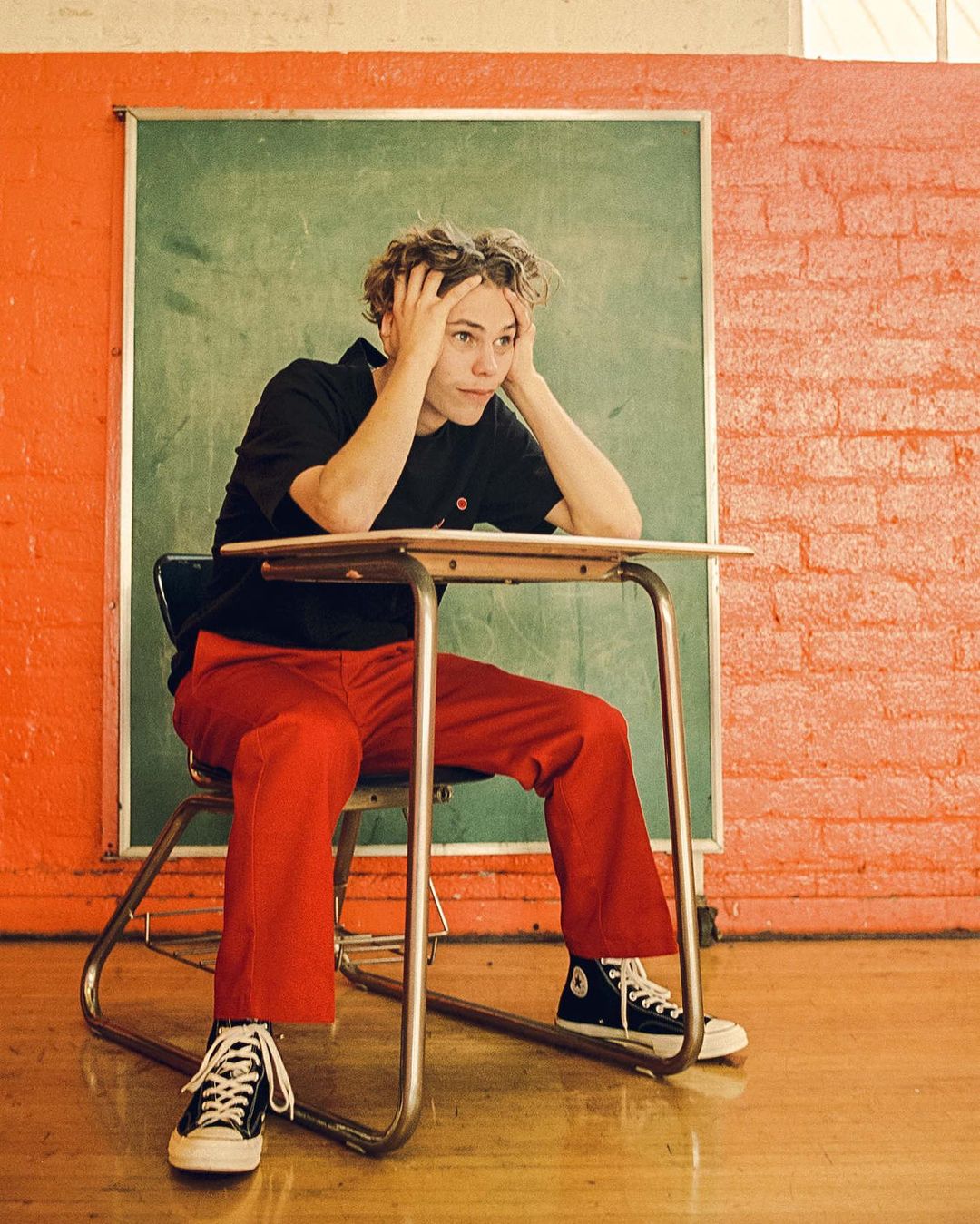 Young adult male with curly hair sits at a school desk in a classroom setting, wearing black shirt, red pants, and Converse sneakers.