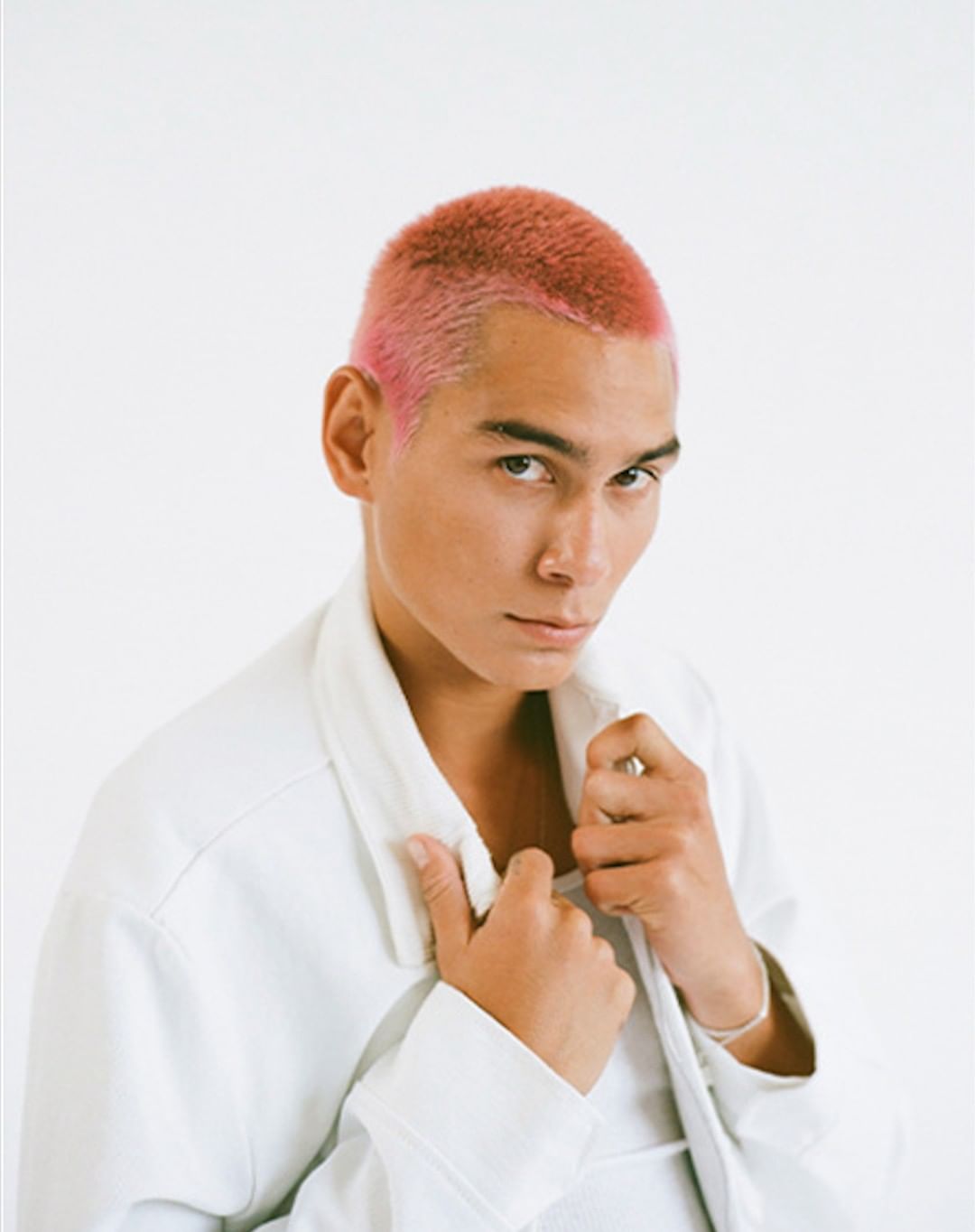 Portrait of adult male with pink buzzcut wearing a white jacket in a photo studio with a white backdrop and medium camera shot.