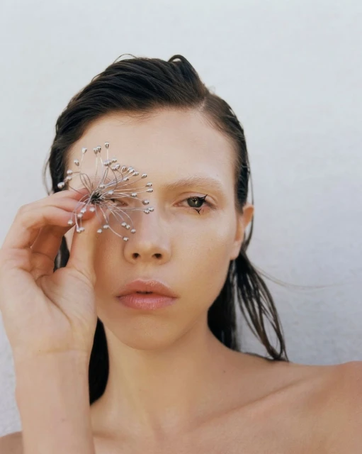 Portrait of woman holding ornamental flower that partially obscures her eye, artistic eye makeup, wet hair, minimal background.