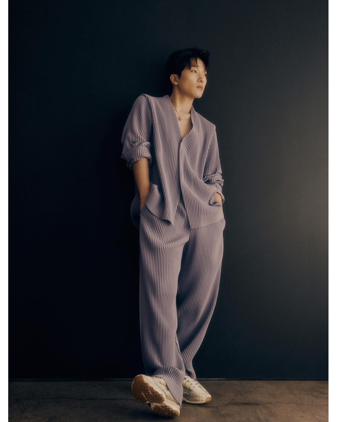 Man in lavender pleated shirt and pants stands against a dark wall, looking away from camera with hands in pockets and sneakers on.