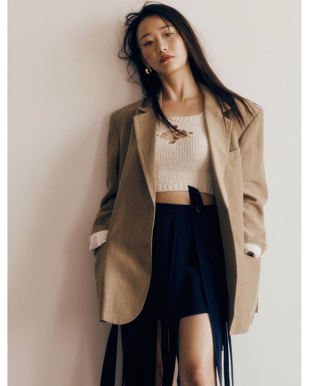 Portrait of a woman with long brown hair, wearing a tan blazer, a cream top, and a black skirt against an off-white background.