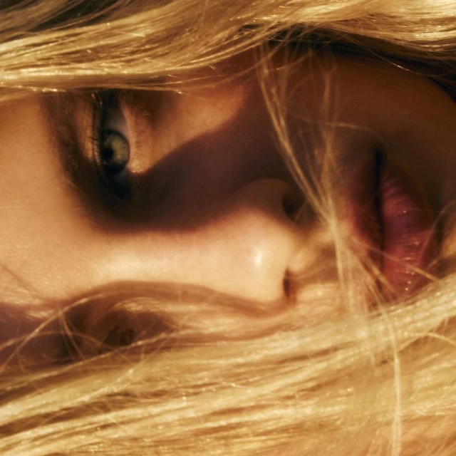 Close-up portrait of a blonde woman with light skin featuring a single visible blue eye and a portion of her face and flowing hair.