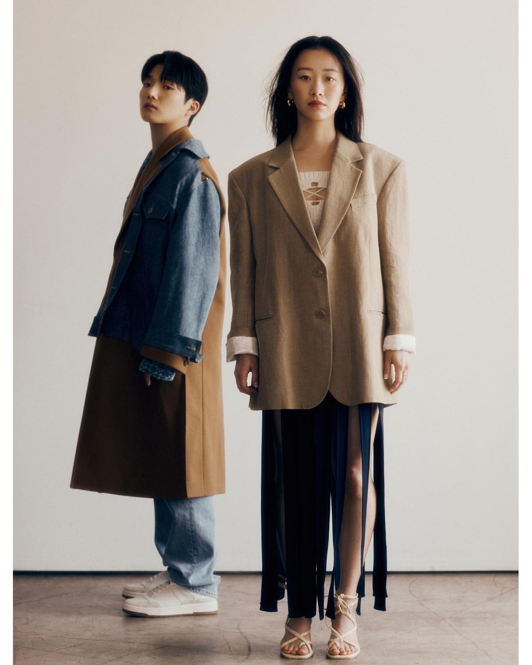 Two fashion models pose in a photo studio. The woman is in a beige blazer, while the man wears a denim jacket and brown coat.