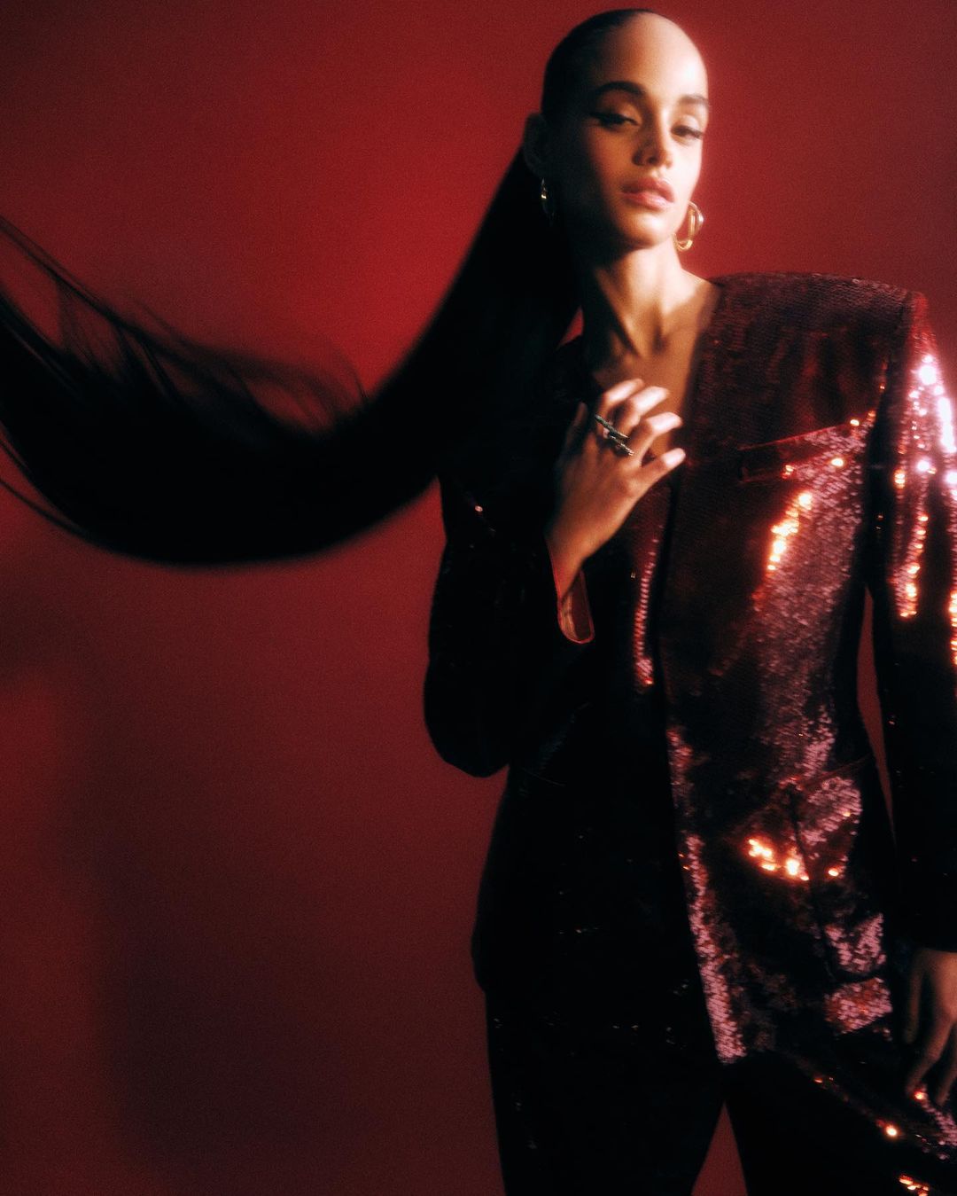 Elegant woman in sequin blazer poses against red backdrop, her long dark hair cascading dramatically, showcasing high-fashion and sophistication.