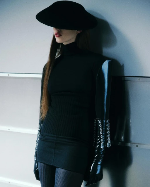 Fashion shot of a woman in a black turtleneck, black gloves, and a wide-brimmed hat against a neutral background.