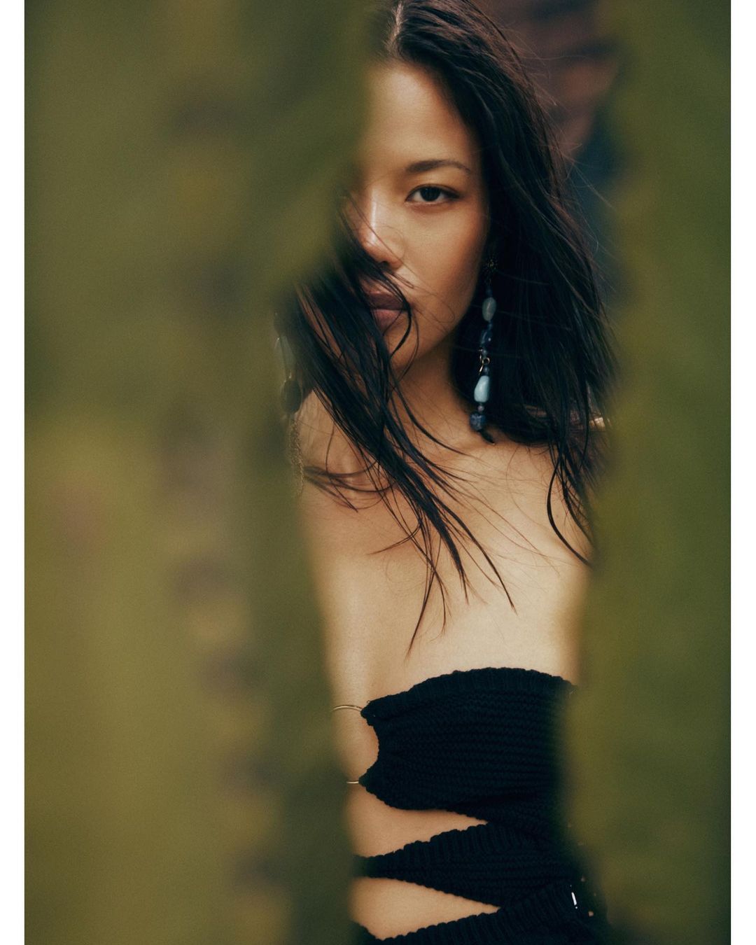 Portrait of a woman with long dark hair wearing a black strapless dress and dangle earrings, partially obscured by blurry green foliage.