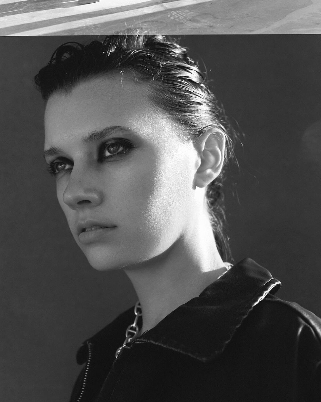 Monochrome portrait of a woman with dark smudged eye makeup wearing a dark jacket and chain necklace by Jeremy Choh