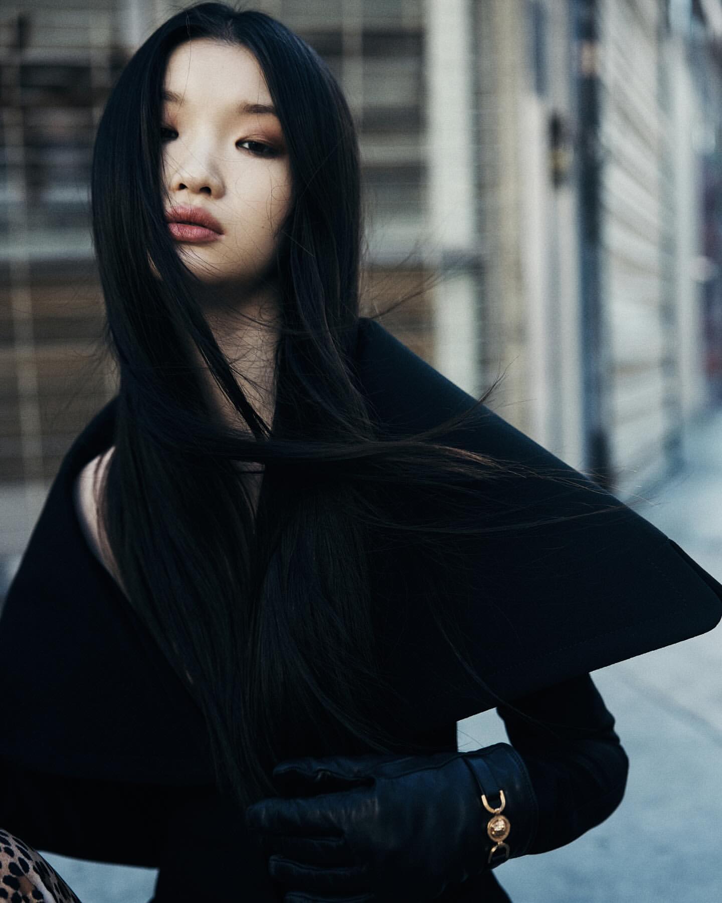 Portrait of a young woman with long dark hair, wearing a black coat and gloves, standing in front of an urban building.