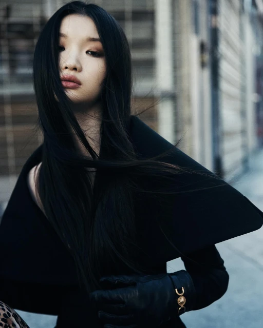 Portrait of a young woman with long dark hair, wearing a black coat and gloves, standing in front of an urban building.