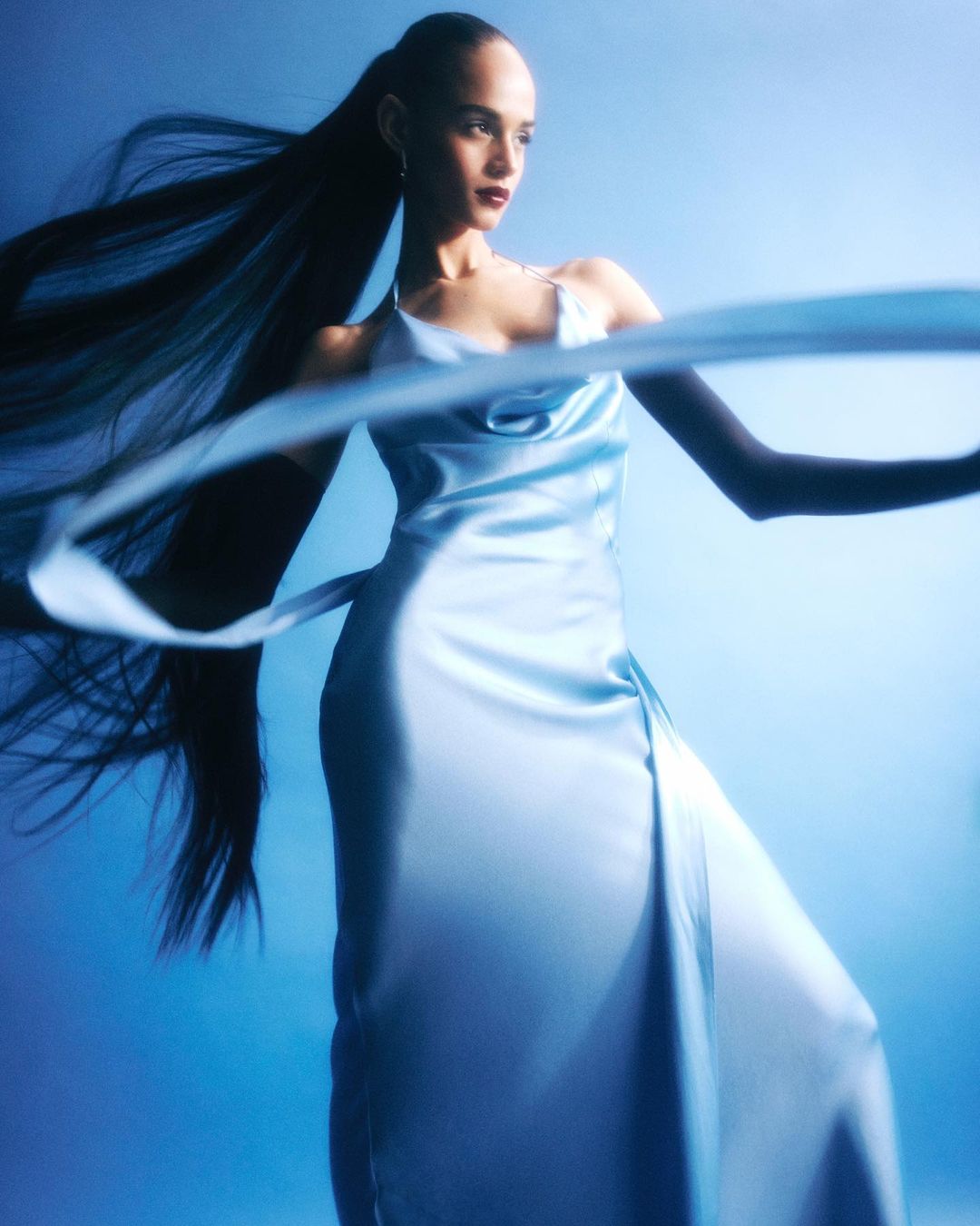 A woman with long black hair wearing a light blue dress, posing gracefully against a soft blue background, creating an ethereal and elegant visual.