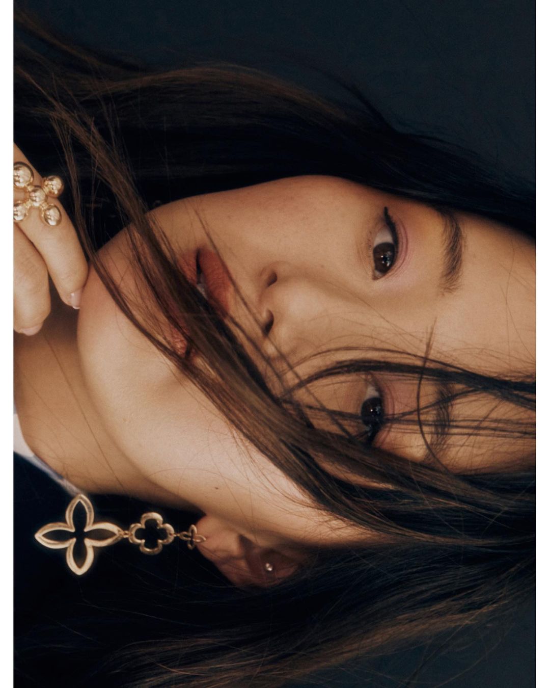 Close-up studio portrait of a woman with long brown hair, wearing gold flower earrings and a ring, against a dark background.