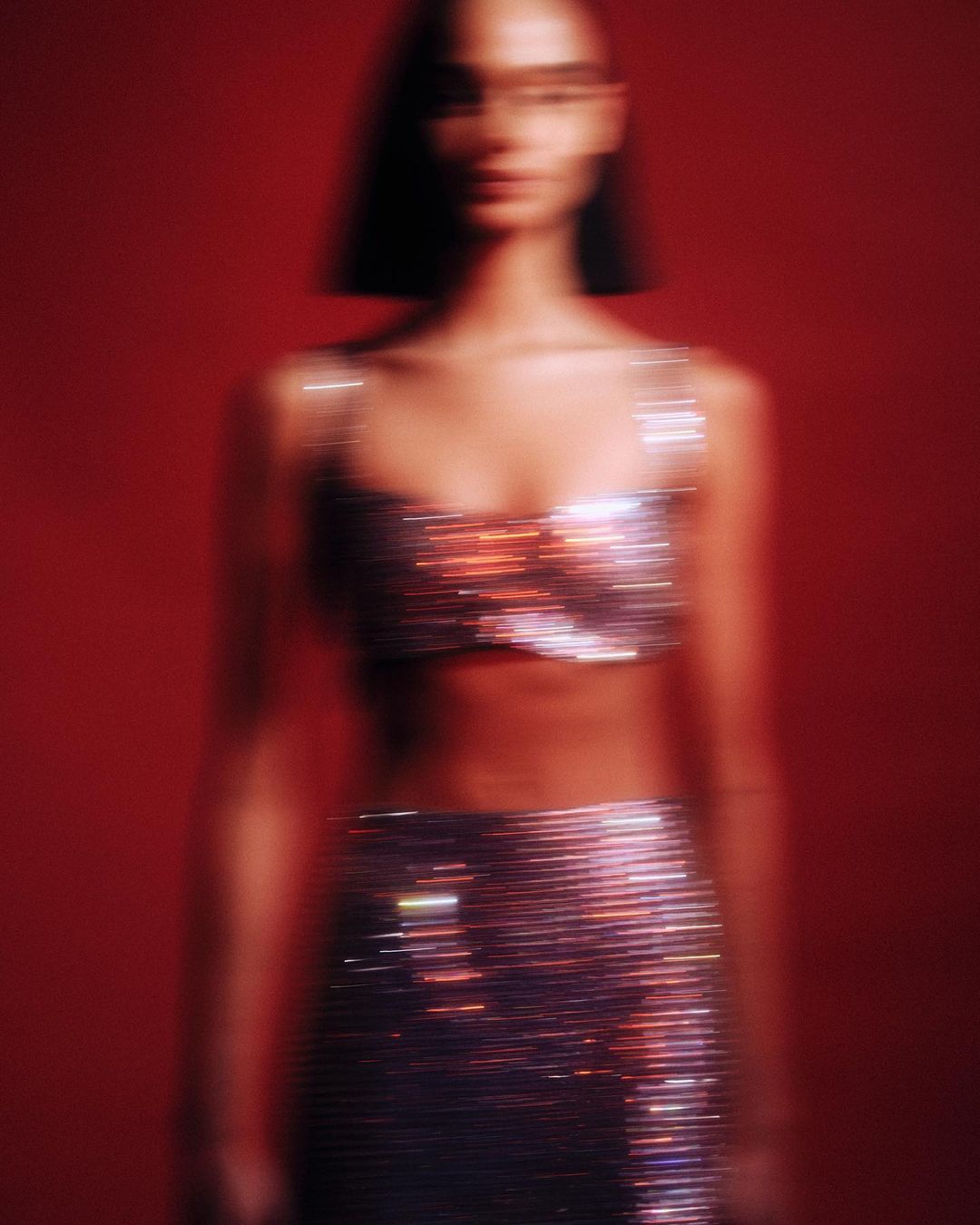 Blurred fashion shot of woman wearing shiny two-piece outfit against red backdrop with soft lighting and shallow depth of field.