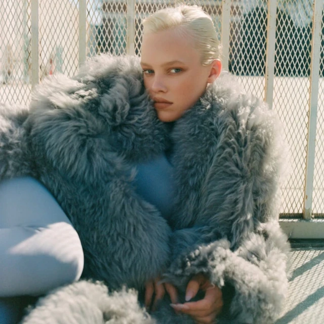Fashion photo of a young adult woman with a short blonde haircut posing while wearing a large fur coat outdoors on a sunny day.