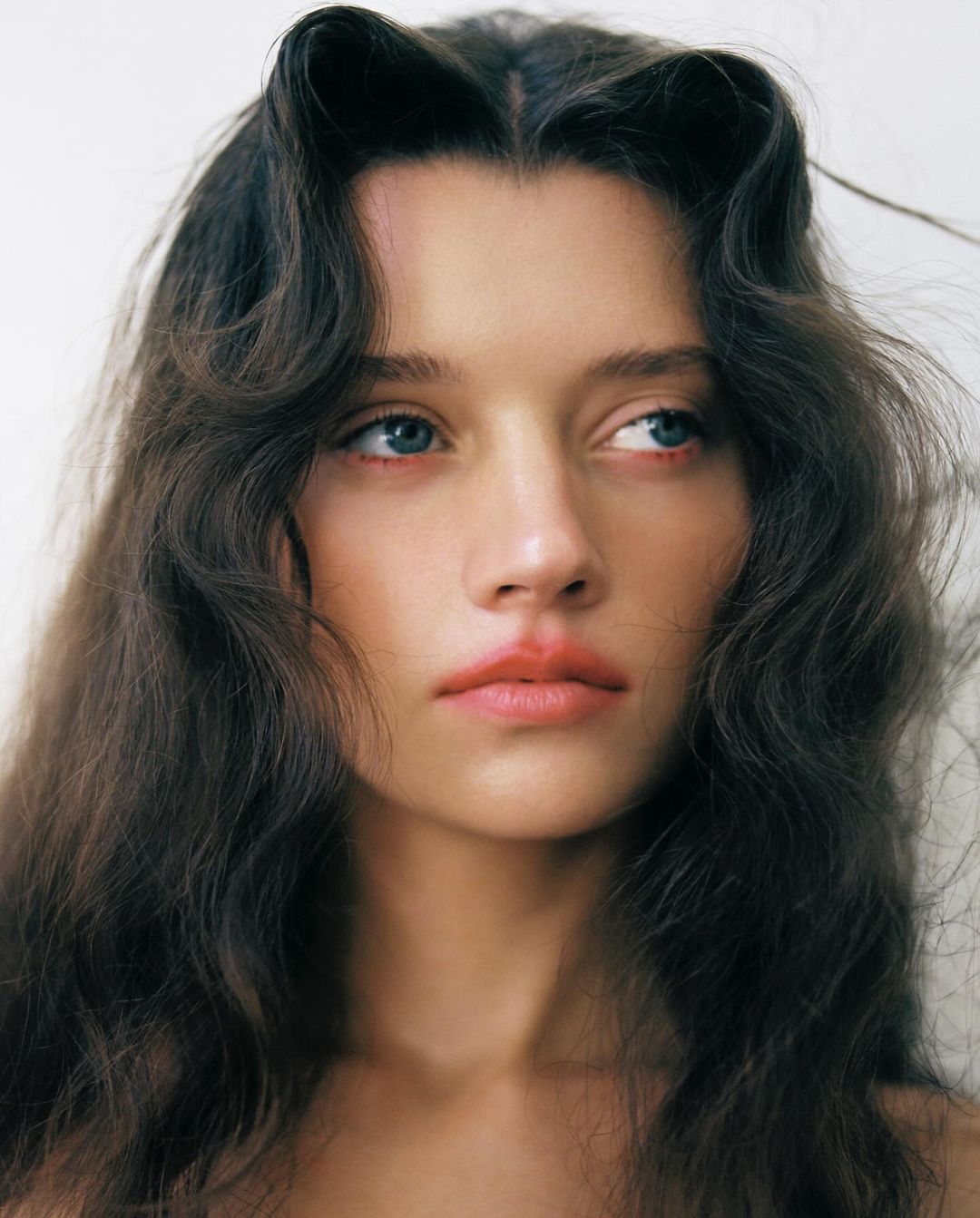 Close-up portrait of a young woman with dark wavy hair styled with faux bangs, light skin, and blue eyes against a white background.