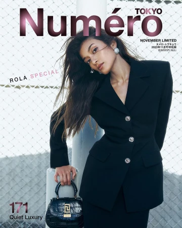 A woman wearing a structured black blazer holding a dark handbag in front of a chain-link fence, captured by Jeremy Choh.