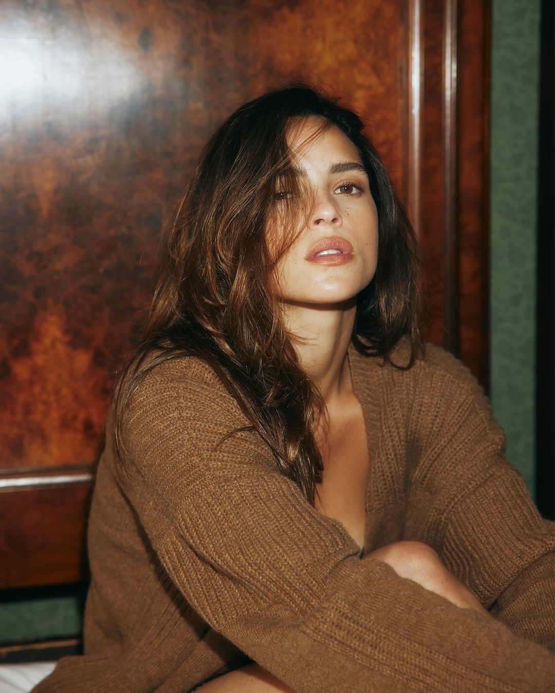 Portrait of a woman with long brown hair wearing a brown cardigan, posing indoors against a dark wooden panel. Beauty and fashion shot.