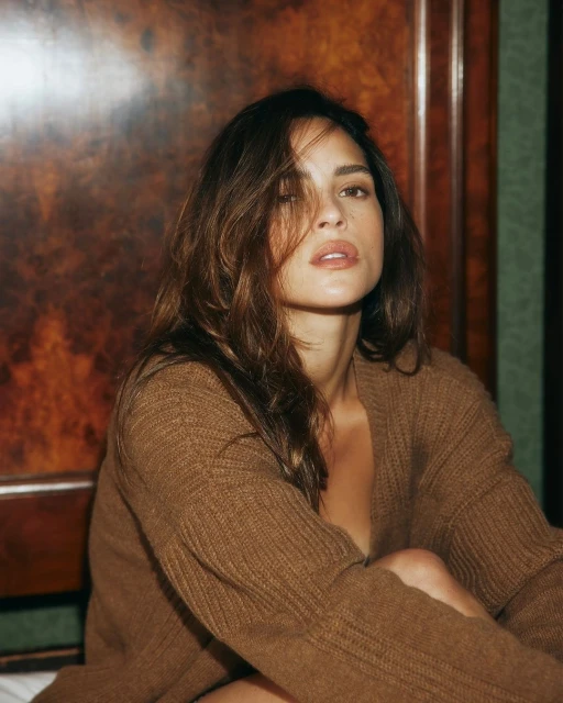 Portrait of a woman with long brown hair wearing a brown cardigan, posing indoors against a dark wooden panel. Beauty and fashion shot.