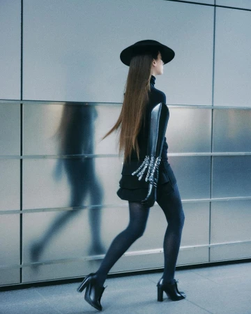 Profile of a long-haired woman wearing a black wide-brim hat, turtleneck, skirt, tights, and heels walking past a reflective wall.