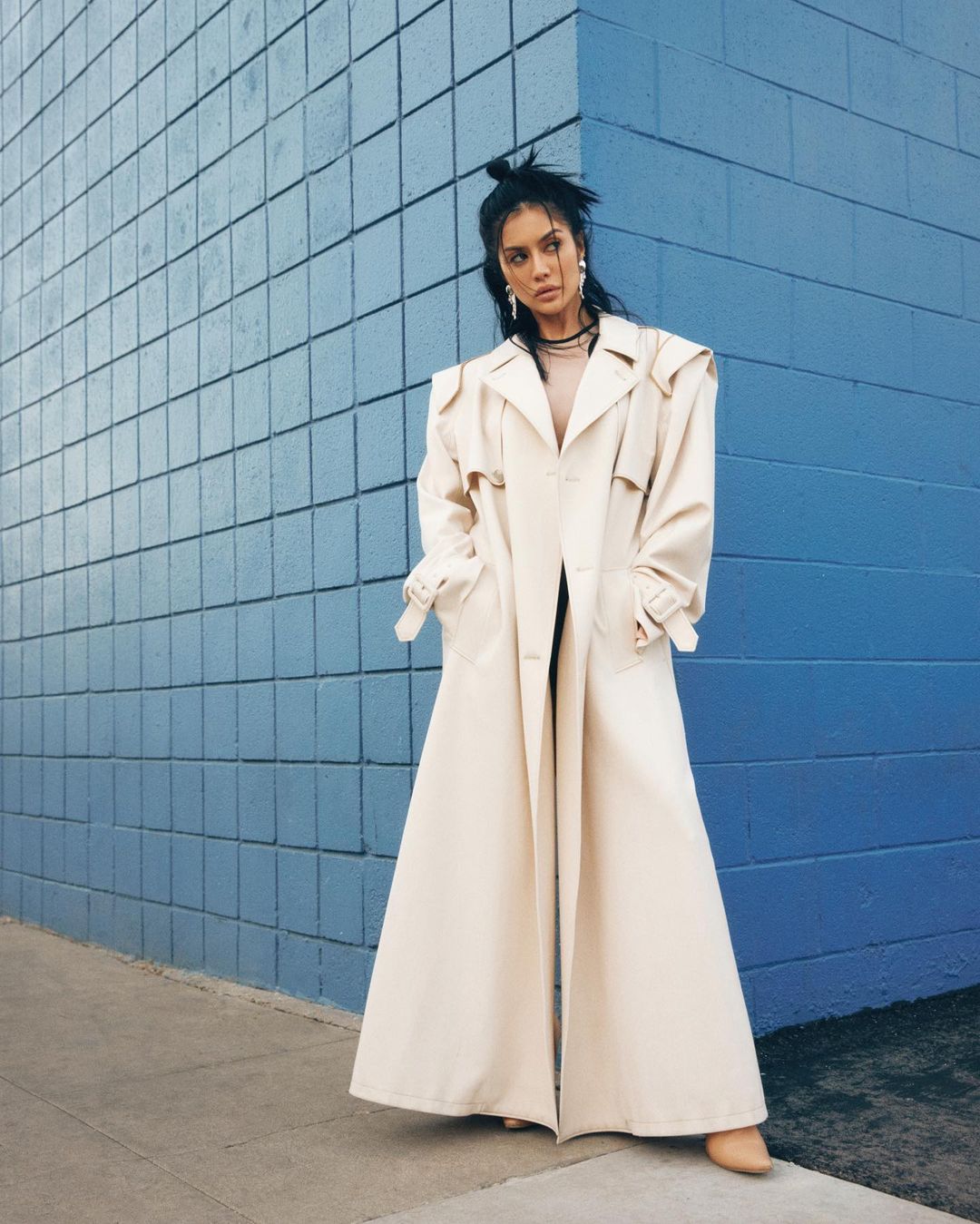 A stylish woman poses against a blue tiled wall wearing a cream-colored trench coat. Her dark hair is in a messy bun and she's wearing jewelry.