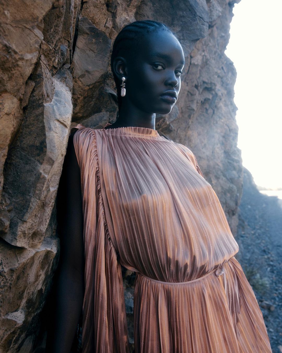 Elegant young woman with dark skin in a pleated dress poses next to a textured rock formation outdoors with soft natural lighting.