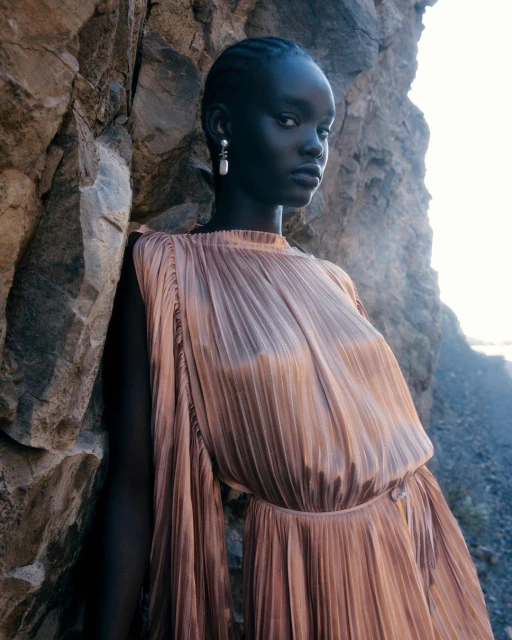 Elegant young woman with dark skin in a pleated dress poses next to a textured rock formation outdoors with soft natural lighting.