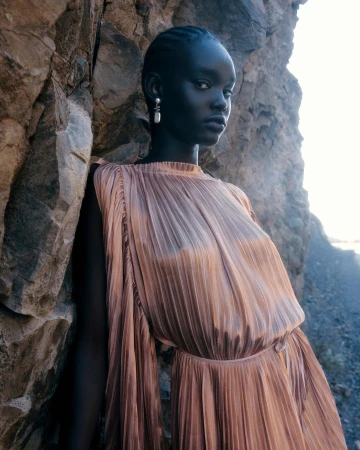 Elegant young woman with dark skin in a pleated dress poses next to a textured rock formation outdoors with soft natural lighting.