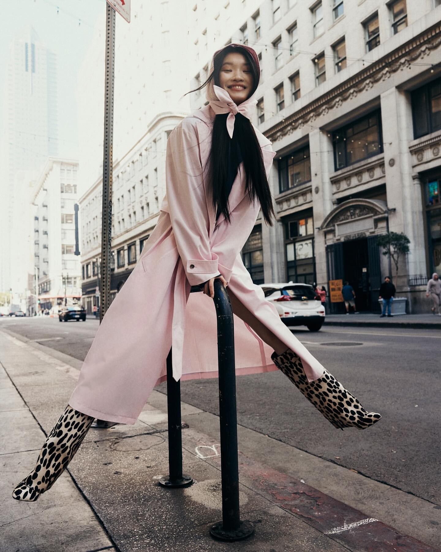 A woman in a pink coat and leopard-print boots poses playfully against a city street backdrop with urban buildings and sidewalks.