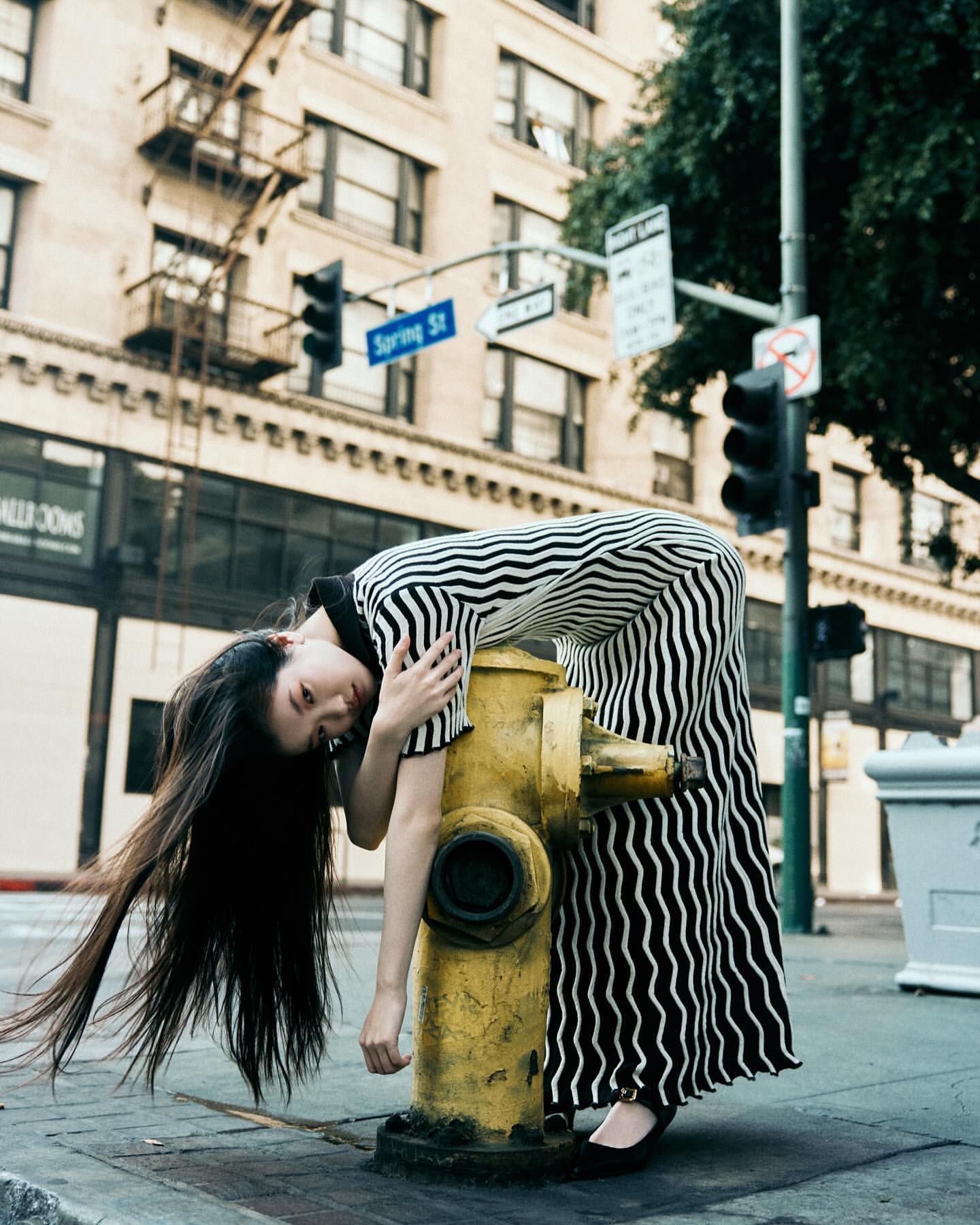 Young Asian woman in black and white striped dress leans on yellow fire hydrant in urban setting with tall buildings and city streets.