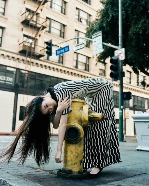 Young Asian woman in black and white striped dress leans on yellow fire hydrant in urban setting with tall buildings and city streets.