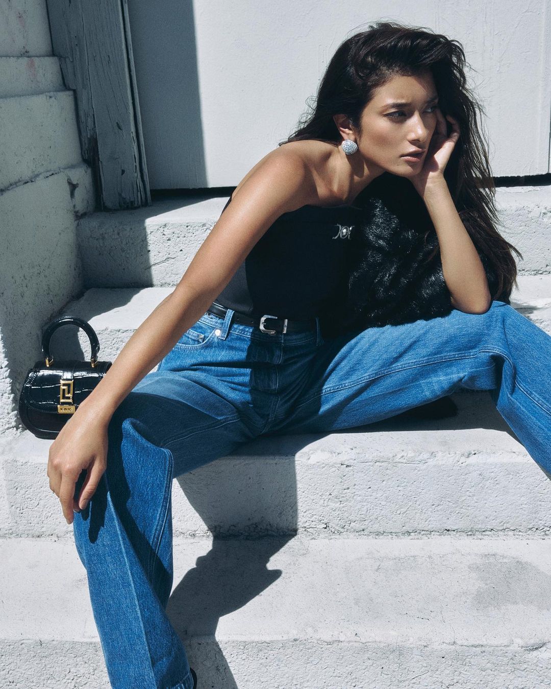 Woman in blue denim jeans and black top sitting on outdoor steps with a black handbag nearby.
