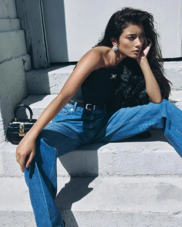 Woman in blue denim jeans and black top sitting on outdoor steps with a black handbag nearby.