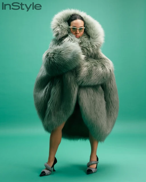 Woman wearing oversized faux fur coat and high heels in studio fashion shot for magazine, set against a teal backdrop.