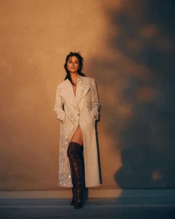Woman in an embroidered light coat and dark thigh-high boots standing against a warm textured wall by Jeremy Choh