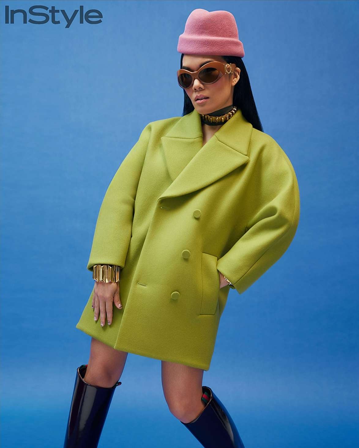 Fashion editorial shot of an Asian woman in a green coat, pink hat, and black boots against a blue backdrop, promoting style and fashion.