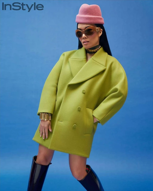 Fashion editorial shot of an Asian woman in a green coat, pink hat, and black boots against a blue backdrop, promoting style and fashion.