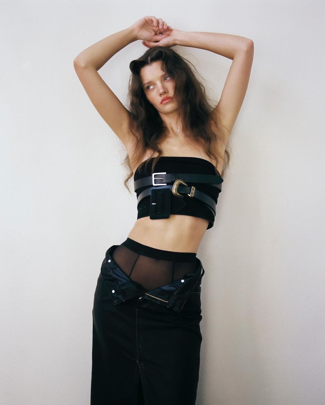 A woman with long wavy hair poses in a black tube top with belts, black skirt, and sheer tights against a white wall in a studio.