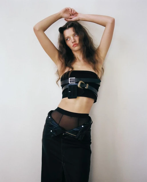 Brunette woman in a black top with belts, open black jeans, and pantyhose, posing indoors against a white wall in a studio setting.