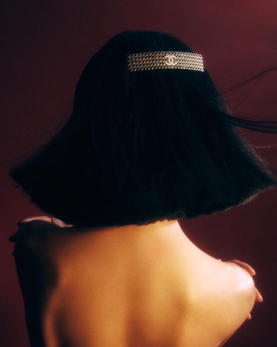 Back view of a woman with short dark hair and bare shoulders, wearing a gold Chanel barrette, against a dark red background.