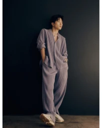 Man wearing a pleated lilac matching set and sneakers standing against a dark wall by Jeremy Choh