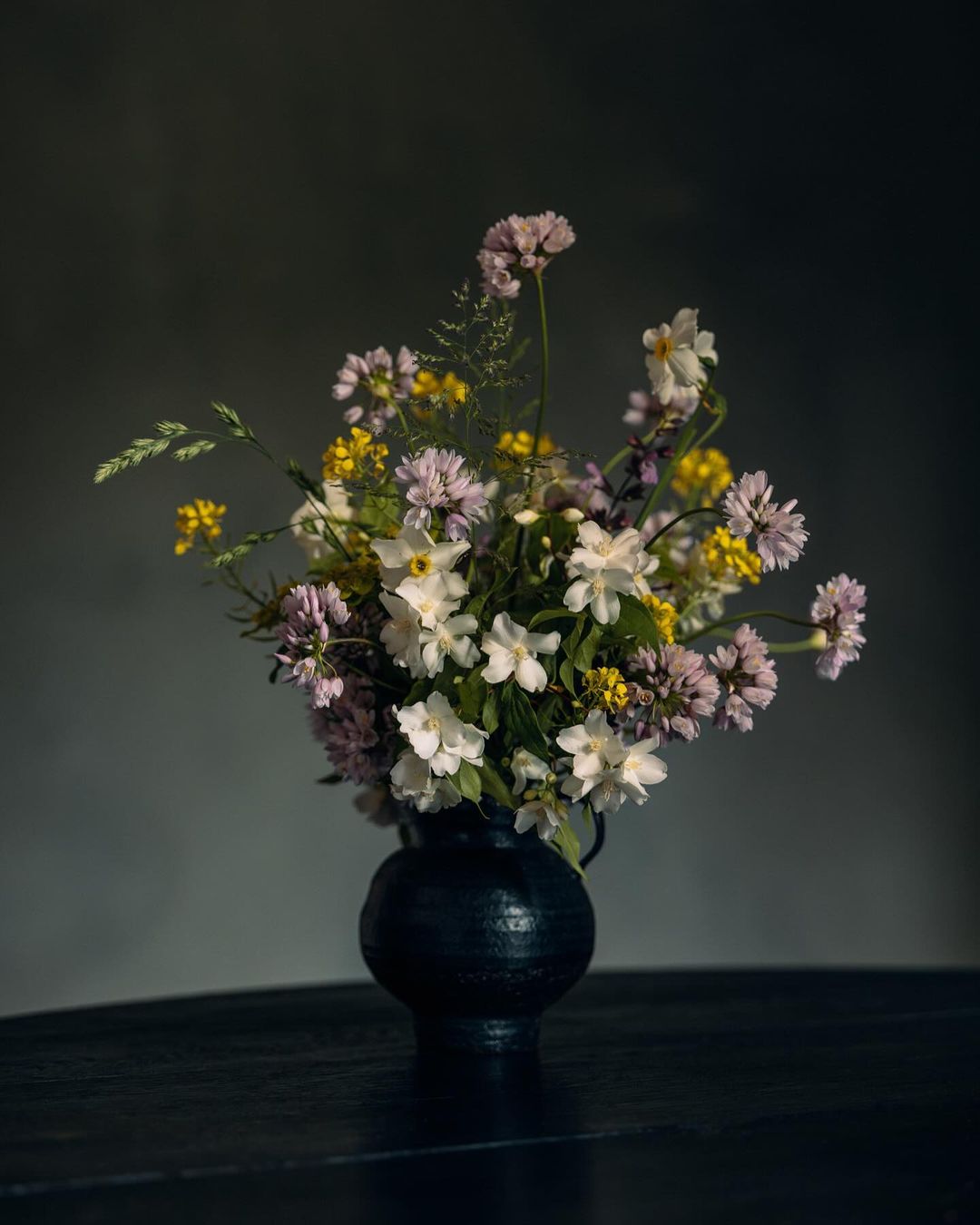 Dramatic floral arrangement of white, pink, and yellow wildflowers in dark, textured vase on dark wood table against grey background.