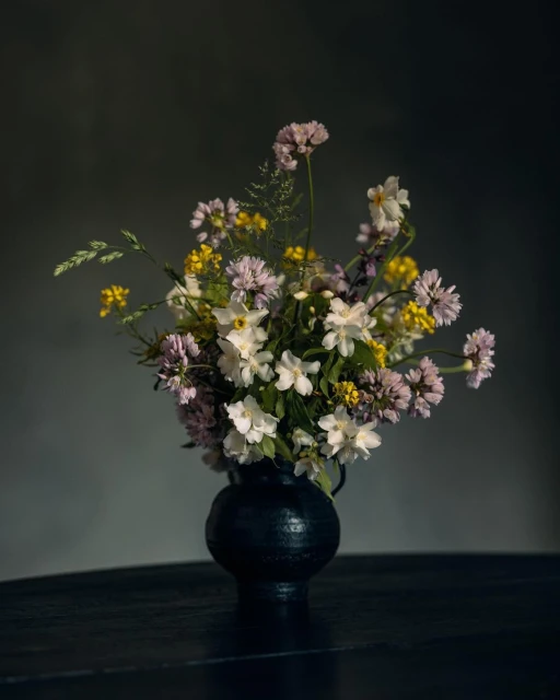 Dramatic floral arrangement of white, pink, and yellow wildflowers in dark, textured vase on dark wood table against grey background.