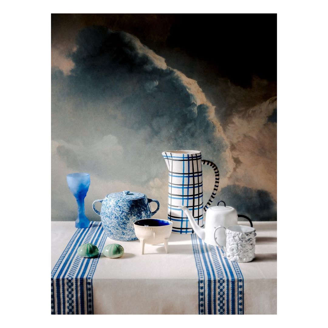 Still life with assorted white and blue ceramic vessels on a white tablecloth set against a dramatic, dark cloudy backdrop.