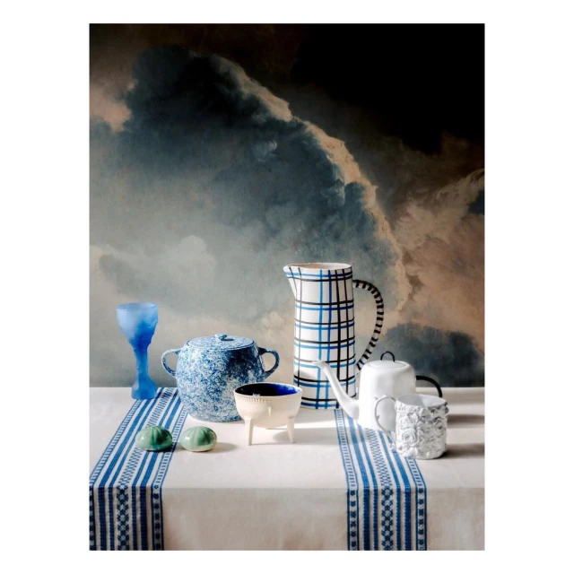 Still life with assorted white and blue ceramic vessels on a white tablecloth set against a dramatic, dark cloudy backdrop.