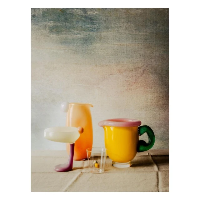 Still life featuring a yellow mug with a green handle, clear glass with a lemon, and peach-colored pitcher against a mottled background.