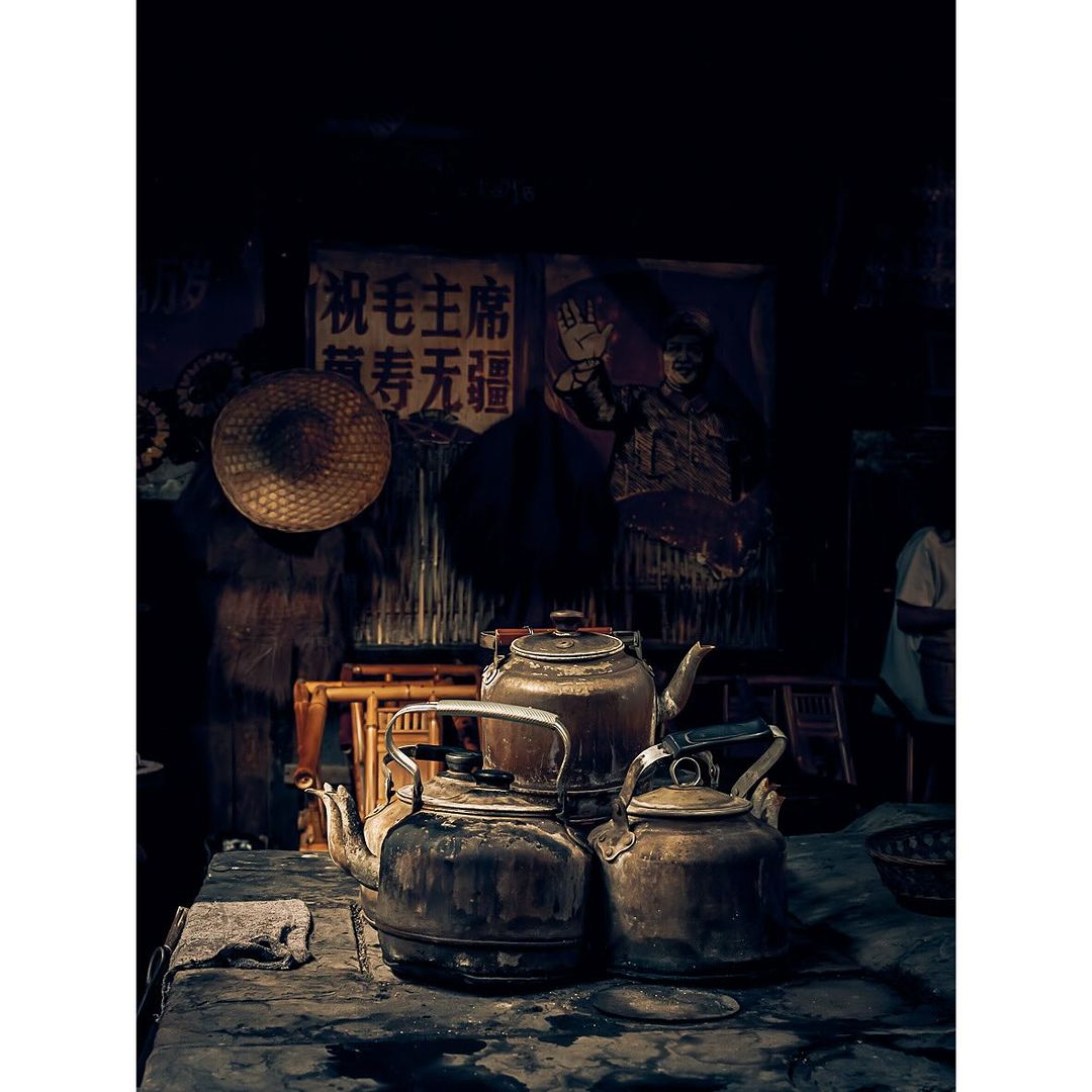 Three rustic teapots sit on a weathered table in dim light, with a woven hat and a political poster in the background.