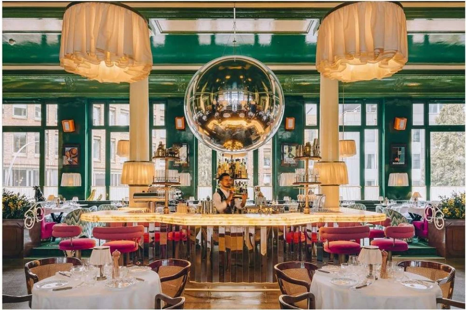 Upscale restaurant interior featuring a mirrored disco ball, seated tables with white linens, and a bartender behind a lighted bar.
